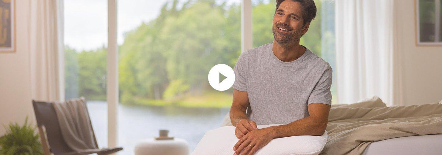 A man set on edge of a bed looking to camera with a play video button overlayed